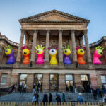A historic building with tall columns displays large, colorful inflatable monster sculptures with big eyes and raised hands on its balcony. People walk and sit on the steps below under a clear, blue sky.