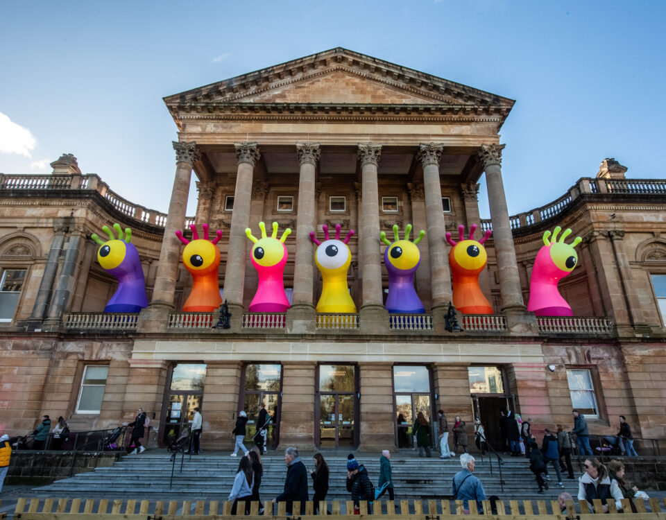 A historic building with tall columns displays large, colorful inflatable monster sculptures with big eyes and raised hands on its balcony. People walk and sit on the steps below under a clear, blue sky.