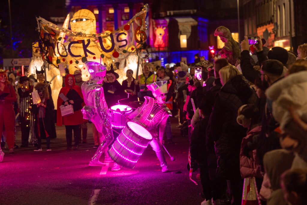 A vibrant street parade at night features two performers in futuristic costumes playing glowing pink drums, while a lively crowd watches. Behind them, a large puppet and a banner with bold, colorful letters are visible.