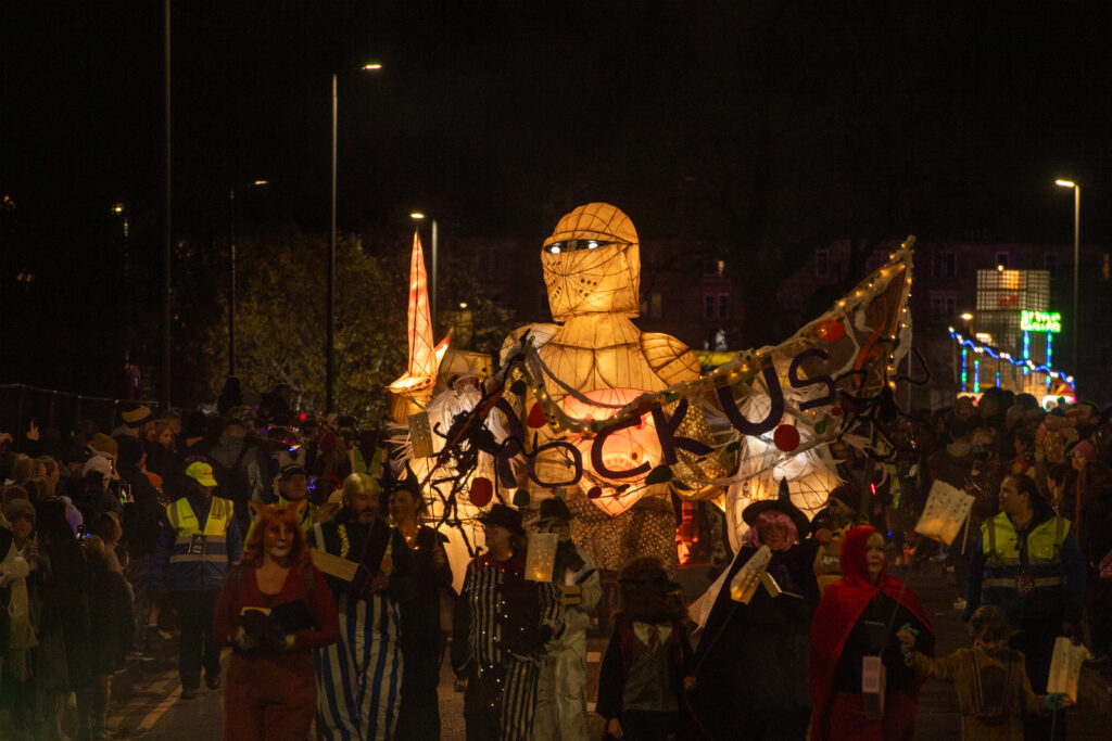 A nighttime parade featuring a large illuminated puppet of a knight surrounded by people in costumes, some carrying lanterns and banners, with bright streetlights and onlookers in the background.
