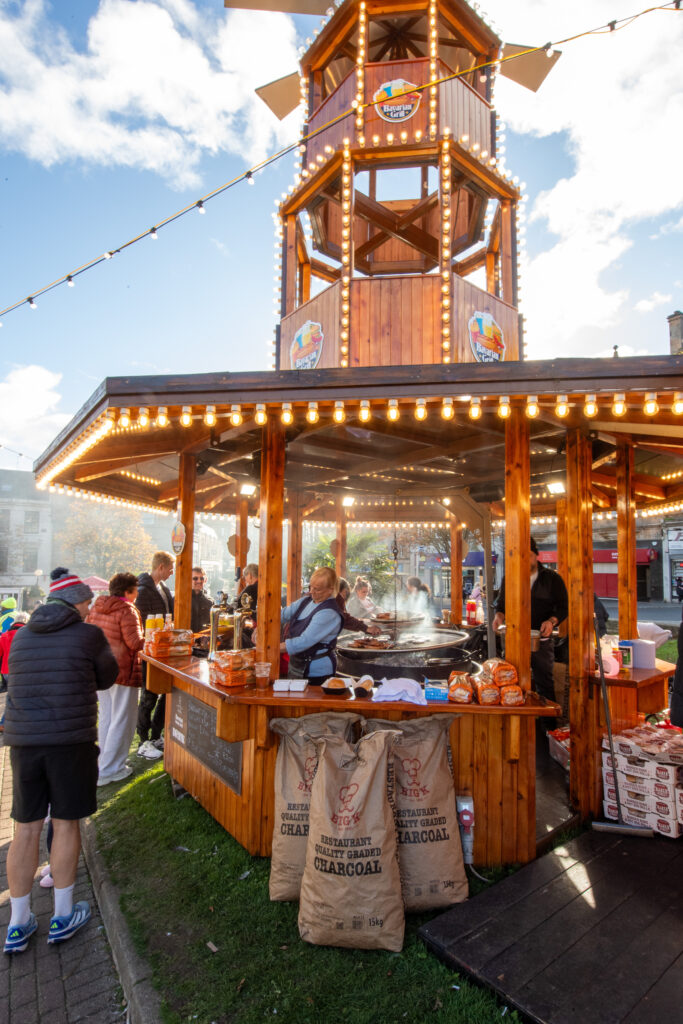 A wooden outdoor food stall with string lights and a tower serves grilled food. People are gathered around, and bags of charcoal are stacked in front. Smoke rises from the grill on a bright day.