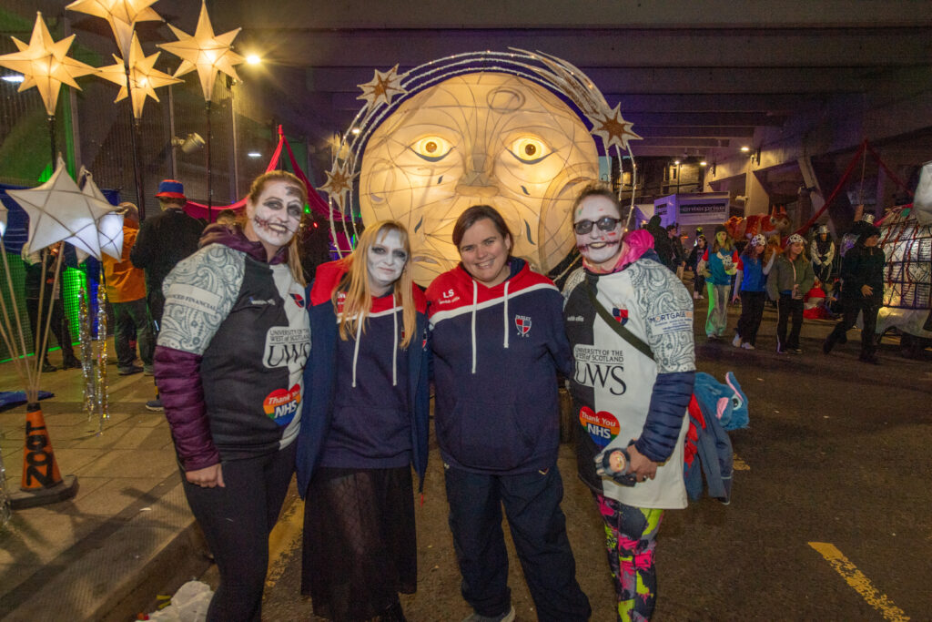 Four people, some with painted faces, pose together at a vibrant nighttime parade with a large illuminated moon face and star lanterns in the background. Crowds and colorful lights fill the festive street scene.