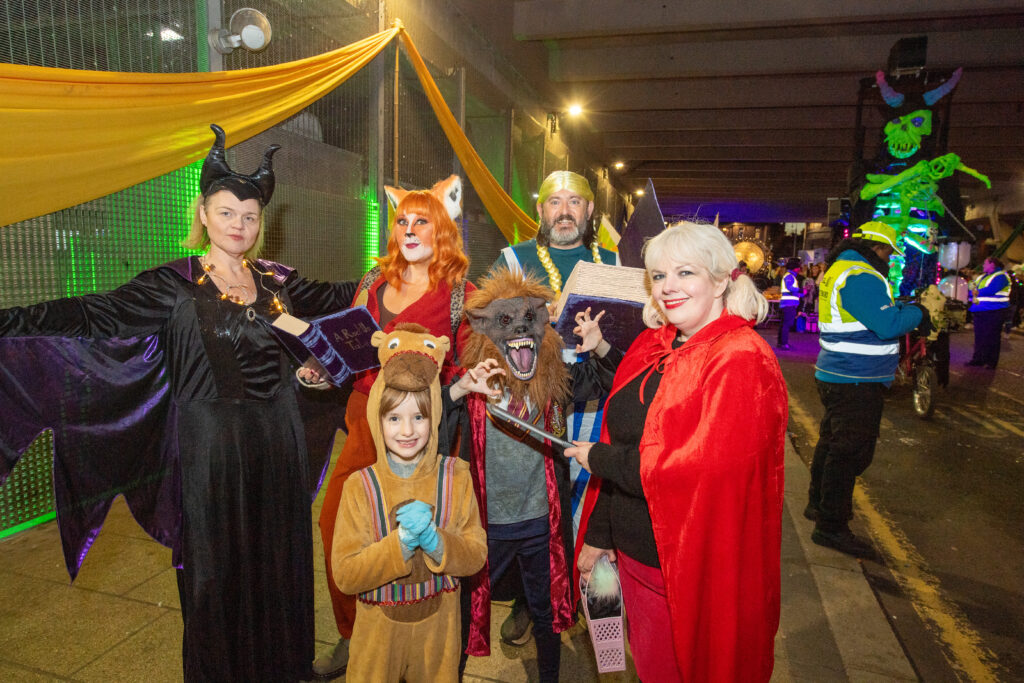 A group of people in colorful fairy tale and animal costumes pose together at a festive nighttime event under streetlights and decorations, with volunteers and a glowing green puppet in the background.