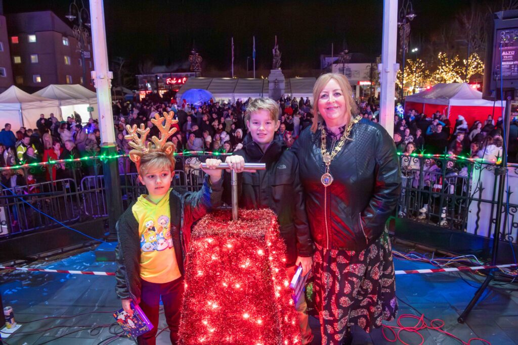 A woman and two boys stand onstage by a red box covered in Christmas lights, with one boy wearing reindeer antlers. A large crowd and festive lights are visible in the background at night.