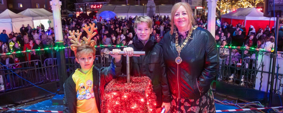 A woman and two boys stand onstage by a red box covered in Christmas lights, with one boy wearing reindeer antlers. A large crowd and festive lights are visible in the background at night.