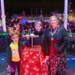 A woman and two boys stand onstage by a red box covered in Christmas lights, with one boy wearing reindeer antlers. A large crowd and festive lights are visible in the background at night.