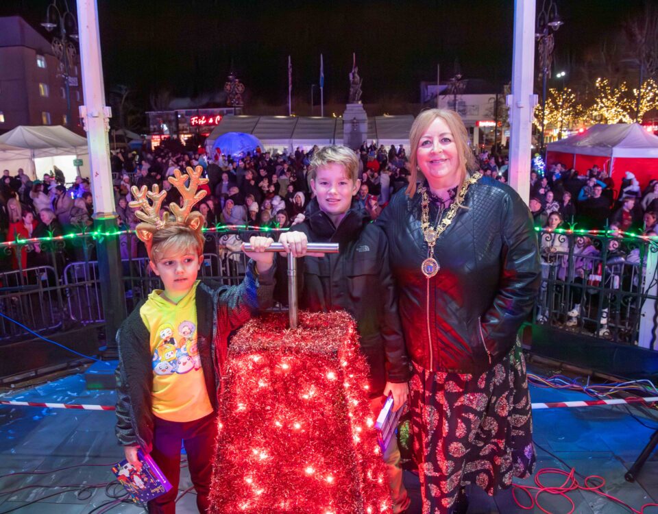 A woman and two boys stand onstage by a red box covered in Christmas lights, with one boy wearing reindeer antlers. A large crowd and festive lights are visible in the background at night.