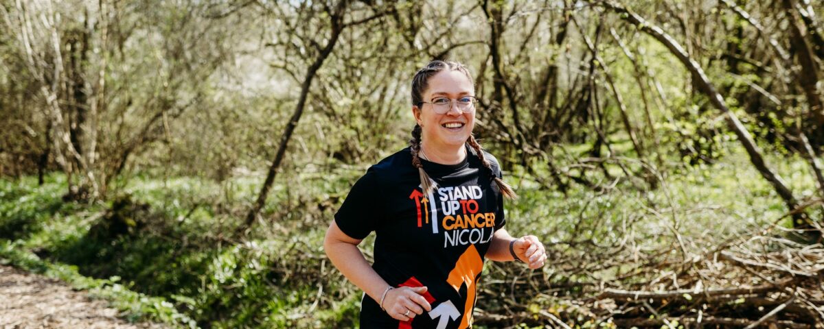 A smiling woman with glasses and braided hair jogs on a wooded trail, wearing a black "Stand Up To Cancer Nicole" T-shirt and shorts. Sunlight filters through the trees, and the path is surrounded by greenery.