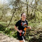 A smiling woman with glasses and braided hair jogs on a wooded trail, wearing a black "Stand Up To Cancer Nicole" T-shirt and shorts. Sunlight filters through the trees, and the path is surrounded by greenery.
