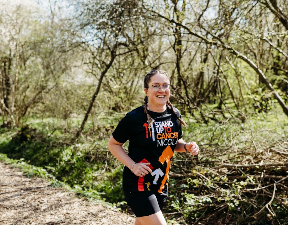 A smiling woman with glasses and braided hair jogs on a wooded trail, wearing a black "Stand Up To Cancer Nicole" T-shirt and shorts. Sunlight filters through the trees, and the path is surrounded by greenery.