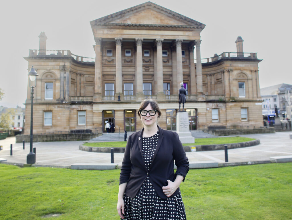 A woman wearing glasses and a black-and-white polka dot dress stands smiling on a lawn in front of a large neoclassical sandstone building with columns and steps.