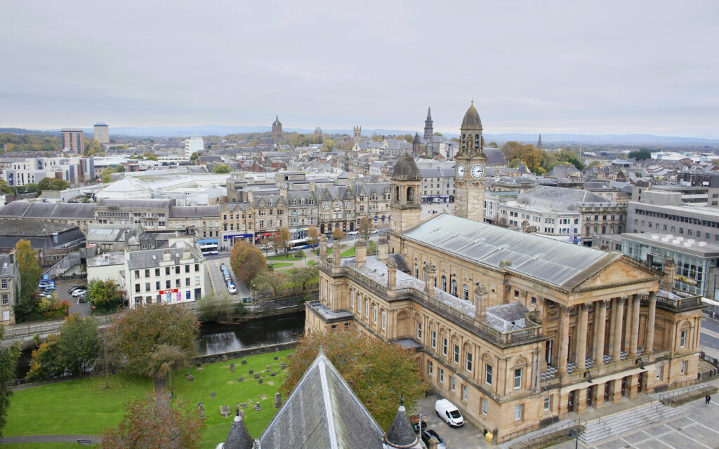 Aerial view of a city featuring a large historic building with columns and a clock tower, surrounded by streets, trees, and other stone buildings under a cloudy sky.
