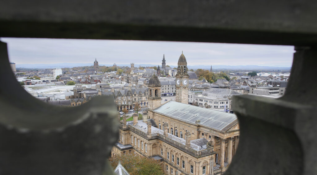 View of a historic cityscape with stone buildings, clock towers, and rooftops, seen through an opening in a stone structure on an overcast day.