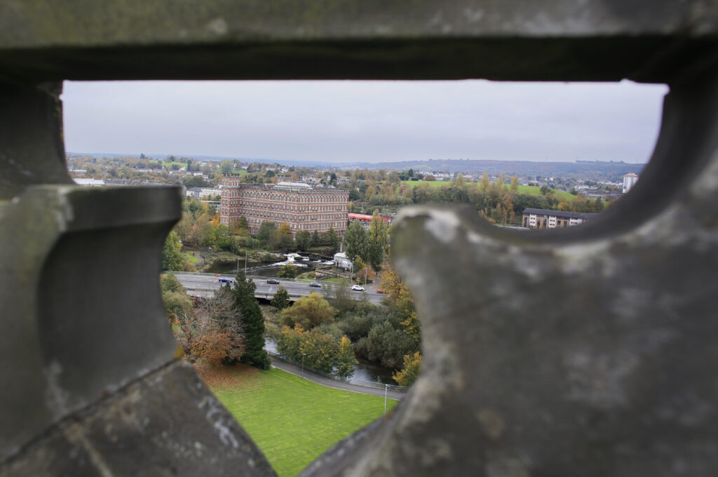 View of a multi-story brick building, trees, and roads in a cityscape, seen through a small opening in a stone wall or structure, with cloudy skies above.