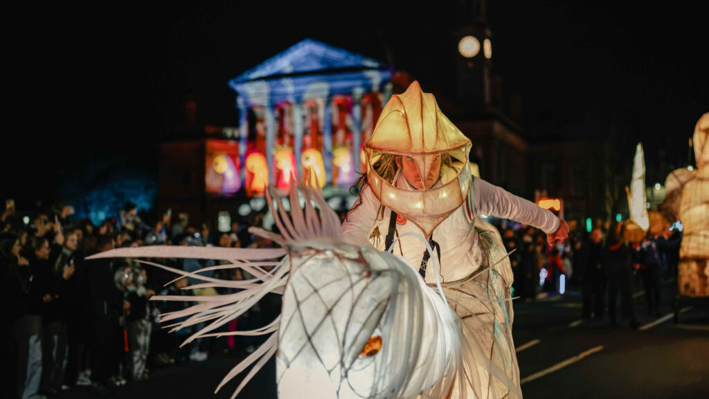 A performer in a luminous sea creature costume rides a similarly styled puppet horse during a nighttime parade, with a brightly lit building and a crowd watching in the background.