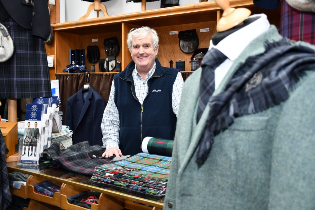 A smiling man stands behind a counter displaying various tartan fabrics in a clothing store, with suits and kilts hanging in the background and a mannequin dressed in a suit and tartan scarf in the foreground.