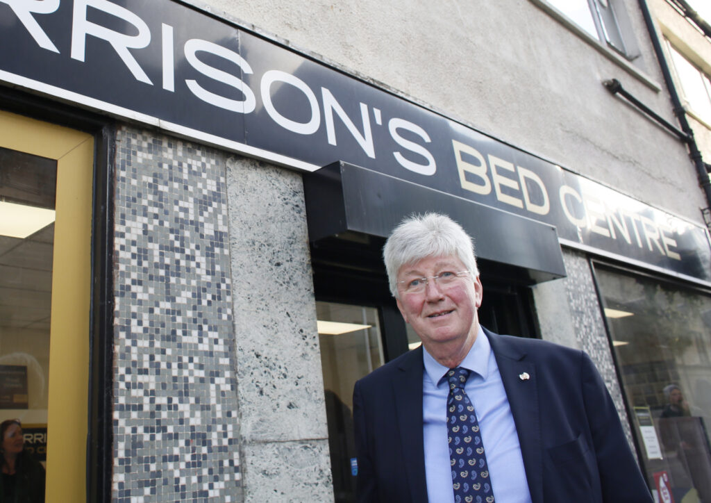 An older man in a suit and tie stands smiling outside a building with a sign reading "RISSON'S BED CENTRE" above the entrance. The exterior features a mosaic tile design beside the door.