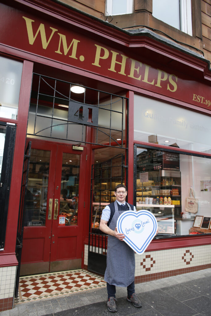 A butcher wearing an apron stands outside W.M. Phelps shop, holding a large heart-shaped “Love Local” sign. The red storefront displays meats and goods through the window.