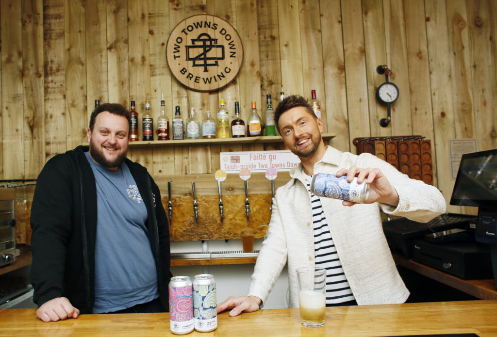 Two smiling men stand behind a wooden bar. One pours a canned drink into a glass while the other stands beside him. Three cans are on the counter, and a “Two Towns Down Brewing” sign hangs on the wall behind them.