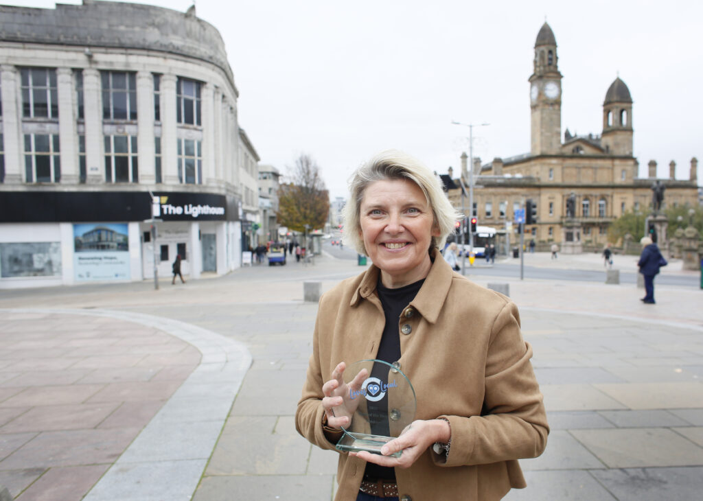 A woman wearing a tan coat smiles while holding a glass award in an urban square with historic buildings and clock towers in the background.
