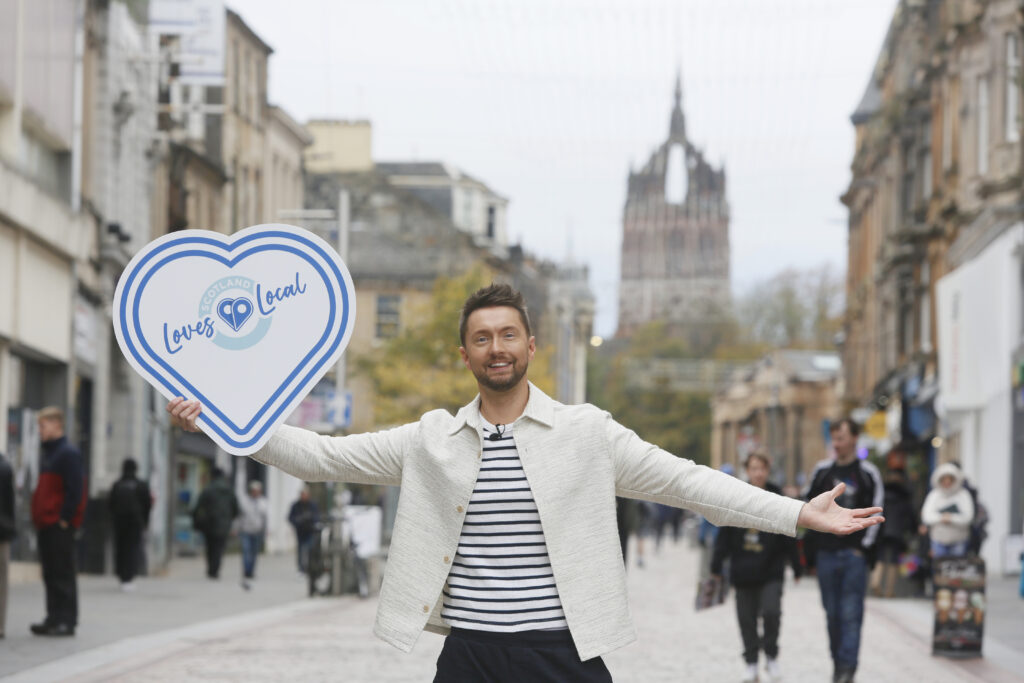 A smiling man stands on a busy city street with arms open, holding a large heart-shaped sign that says "Loves Local." People walk in the background, and a tall, ornate building is visible further down the street.