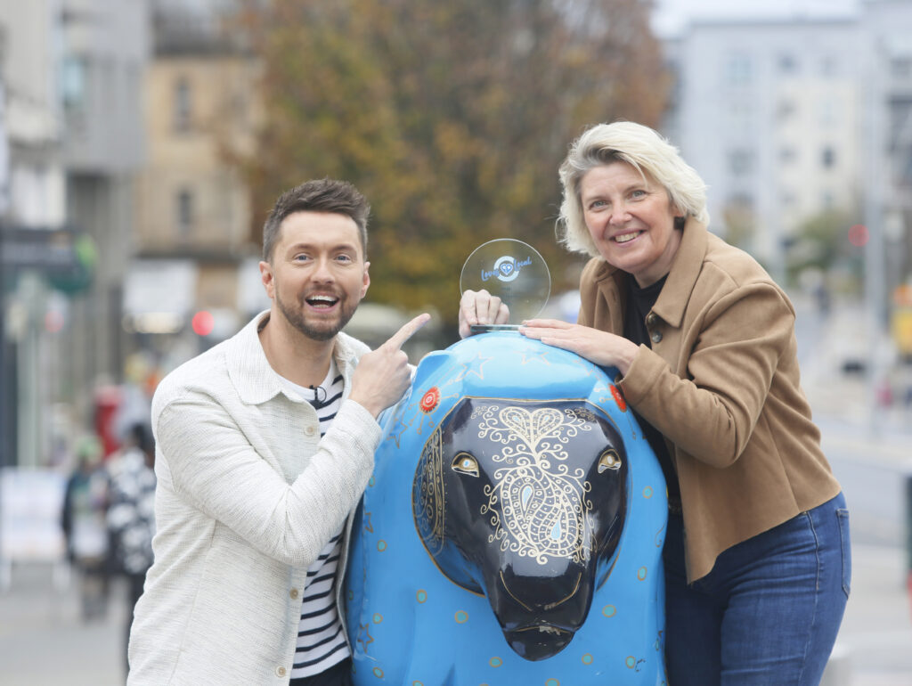 Two smiling people pose outdoors with a blue sheep sculpture decorated with white patterns. One person points at the other, who is holding a small circular award on top of the sculpture. Buildings and trees are visible in the background.