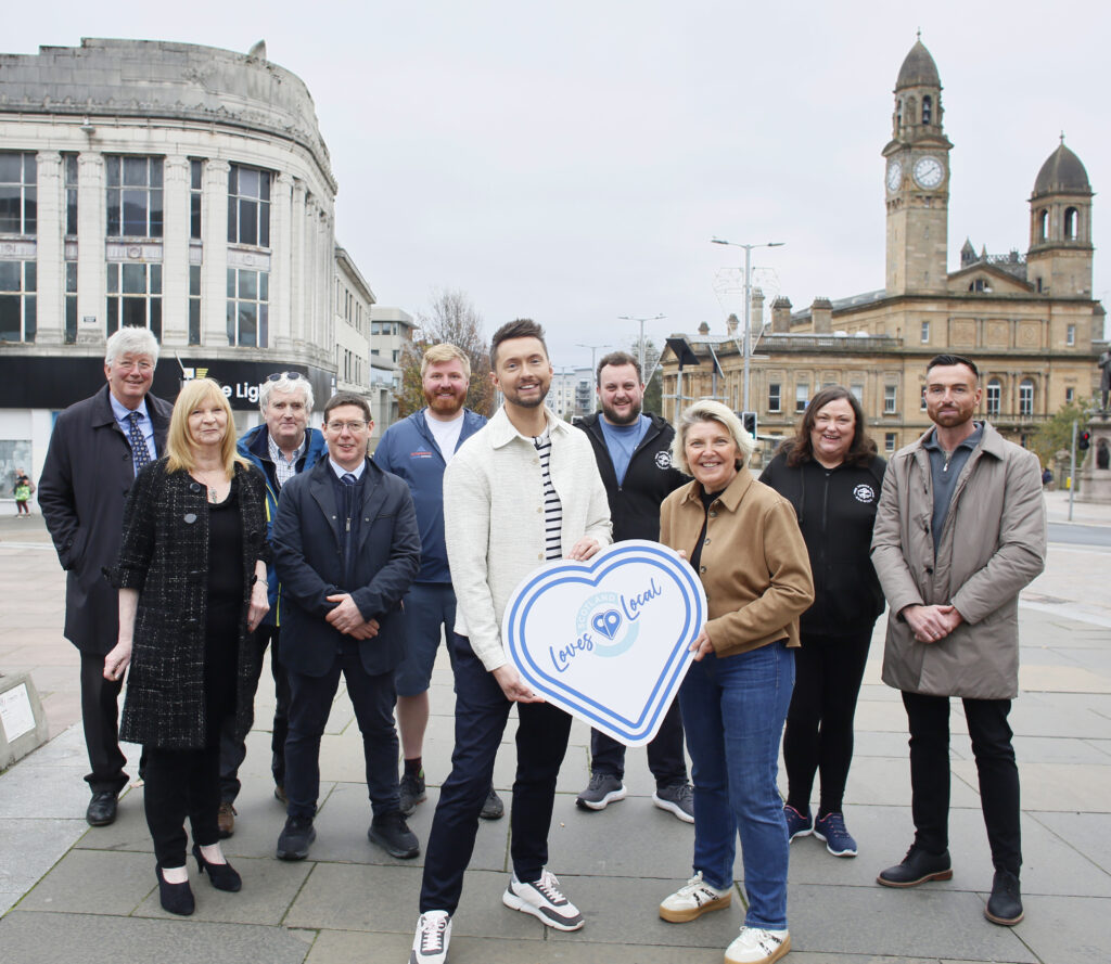 A group of ten people stand together outdoors in a town square, smiling at the camera. Two people in front hold a large blue heart-shaped sign that says "Love Local." Buildings and a clock tower are visible in the background.