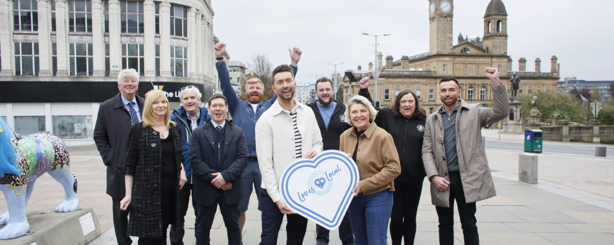 A group of people stand and smile outdoors in a city square, with some raising their arms in celebration. Two people hold a large sign shaped like a heart with "Love Local" written on it. Buildings and a clock tower are in the background.