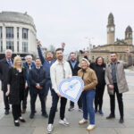 A group of people stand and smile outdoors in a city square, with some raising their arms in celebration. Two people hold a large sign shaped like a heart with "Love Local" written on it. Buildings and a clock tower are in the background.