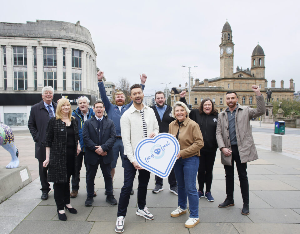 A group of people stand and smile outdoors in a city square, with some raising their arms in celebration. Two people hold a large sign shaped like a heart with "Love Local" written on it. Buildings and a clock tower are in the background.