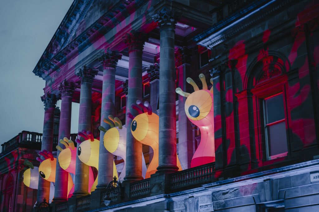 Large illuminated yellow and orange cartoon-like sculptures with big eyes and raised hands are displayed between columns on a grand building, bathed in colorful red and blue lights at dusk.