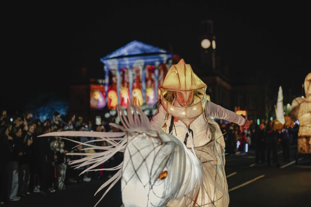 A performer in an illuminated costume resembling a fish or sea creature parades through a crowded street at night, with colorful lights and a historic building in the background.
