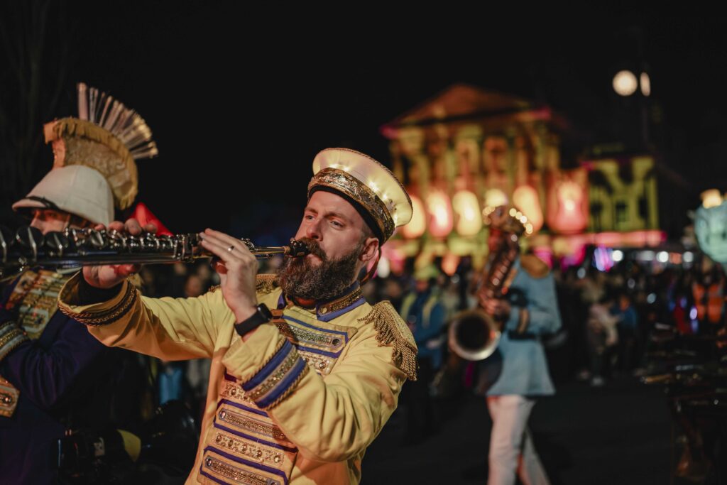 A bearded man in an ornate yellow band uniform plays a clarinet in a lively nighttime street parade, with colorful lights and blurred buildings in the background and other musicians nearby.