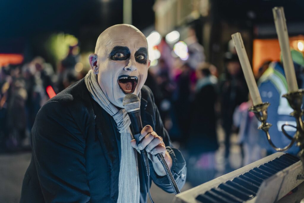 A person with a bald head and dramatic black-and-white face paint sings into a microphone, wearing a dark jacket and scarf, standing next to a keyboard and a candelabra, with a blurred crowd in the background at night.