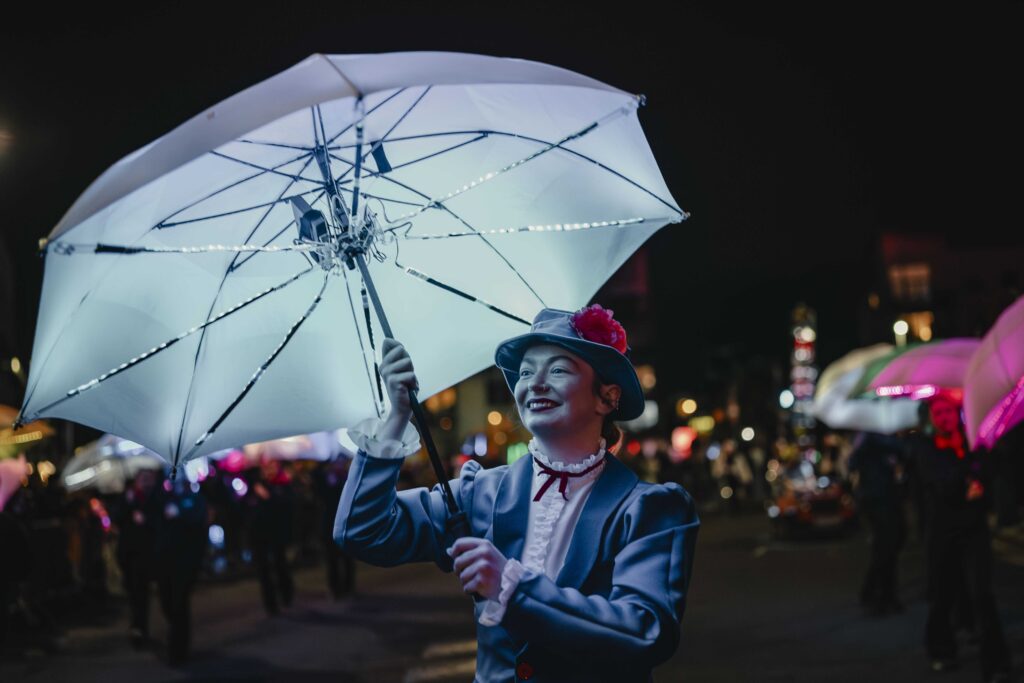 A person dressed in a blue costume and hat with a red flower holds a glowing umbrella at night during a parade, surrounded by others with illuminated umbrellas.