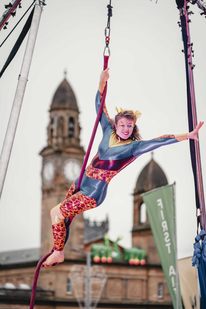 A circus performer in a colorful bodysuit and face paint hangs gracefully from an aerial silk, smiling, with a historic clock tower and green festival banners in the background.