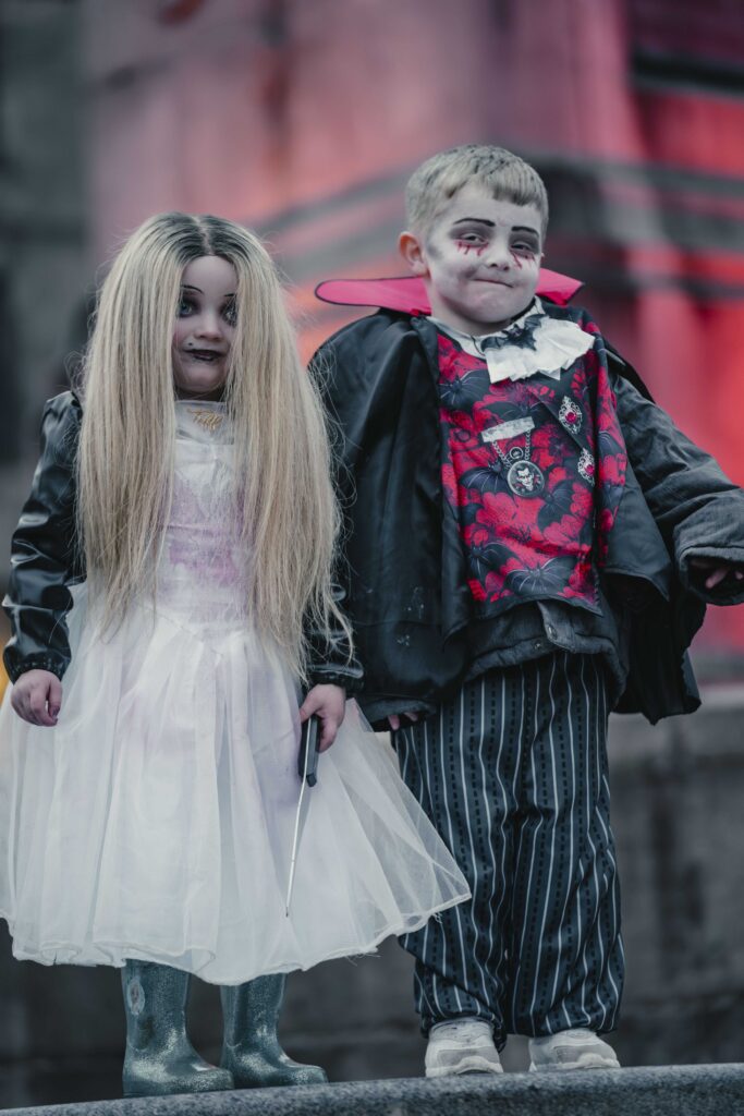 Two children in spooky Halloween costumes: one dressed as a ghostly bride with long blond hair, white dress, and boots, holding a wand; the other as a vampire with face paint, a red vest, cape, and striped pants.