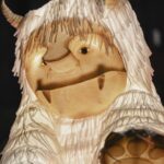 A close-up of a large, illuminated creature with shaggy white fur, two small horns, and a gentle expression, resembling a friendly monster puppet or lantern at a nighttime event.