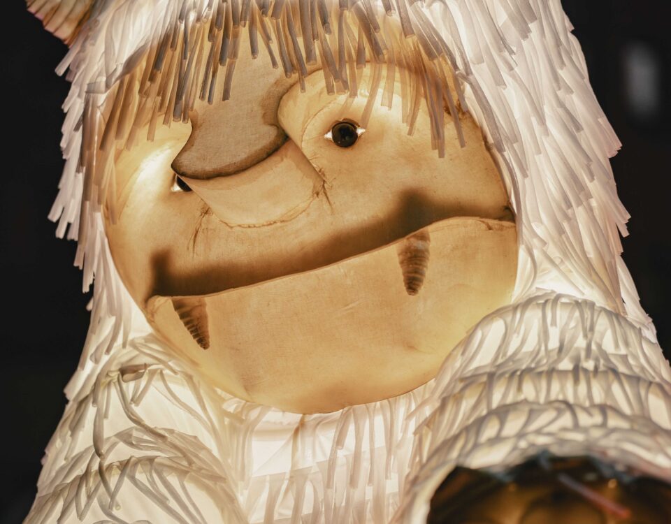 A close-up of a large, illuminated creature with shaggy white fur, two small horns, and a gentle expression, resembling a friendly monster puppet or lantern at a nighttime event.