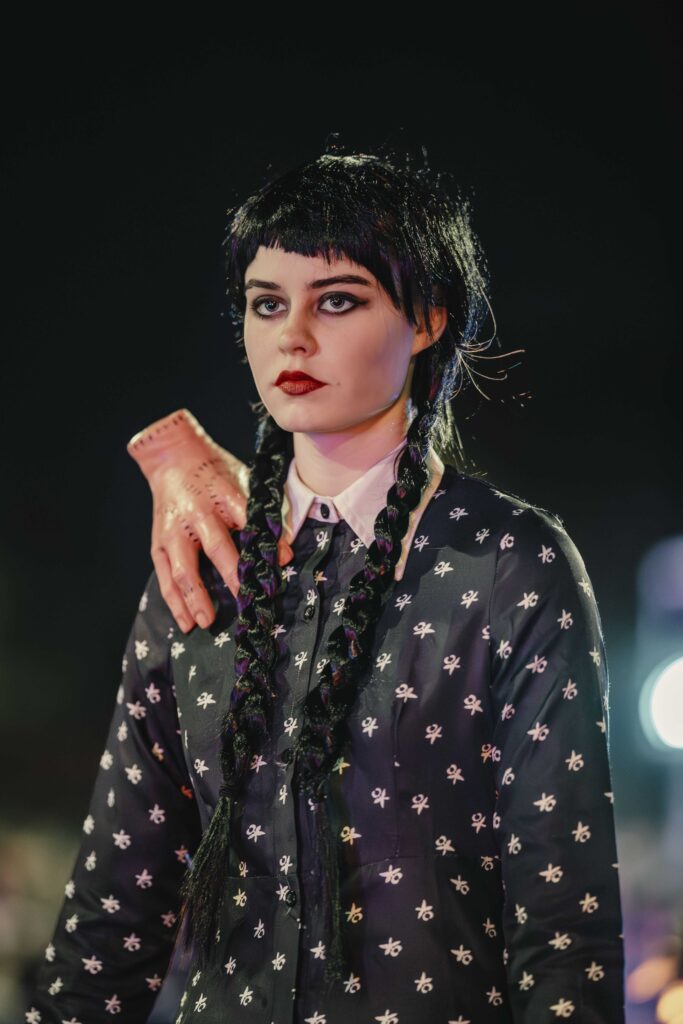 A woman with braided black hair and dramatic makeup wears a black shirt with white patterns. A realistic fake hand rests on her shoulder. The background is dark and blurry with lights.