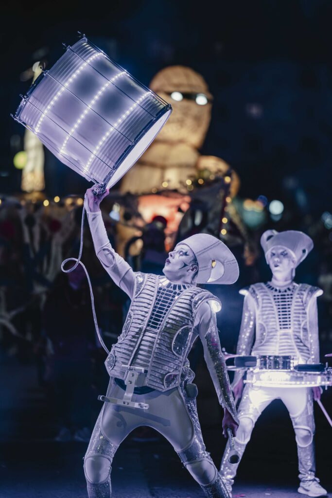 Two performers in futuristic, illuminated costumes play drums at night. One raises a glowing drum above their head. Both wear white face paint and sculpted hats. A large, lit-up figure stands in the background.