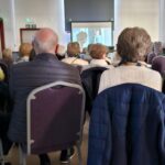 A group of people sit on chairs facing a projection screen in a room, watching a presentation. Most are older adults wearing jackets, and daylight is visible through windows on the right.