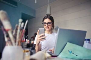 A woman wearing glasses sits at a desk with art supplies, looking at her smartphone and smiling, with an open laptop in front of her in a well-lit room.