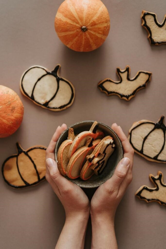 Hands holding a bowl with Halloween-themed cookies shaped like pumpkins and bats, surrounded by more decorated cookies and small pumpkins on a brown surface.