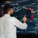 A man in a white shirt points at a large screen displaying colorful financial charts and graphs, analyzing stock market data in a modern trading environment.