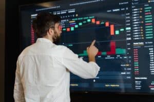 A man in a white shirt points at a large screen displaying colorful financial charts and graphs, analyzing stock market data in a modern trading environment.