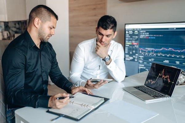 Two men sit at a desk with a laptop and an open notebook, analyzing financial charts displayed on a laptop and a large monitor, appearing focused and engaged in discussion.