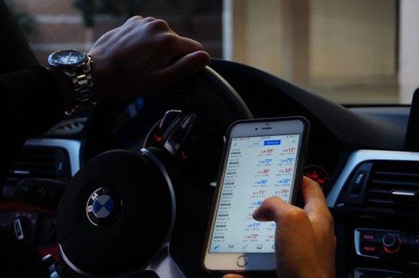 A person is driving a BMW and holding a smartphone with a stock trading app open, displaying various financial data, while wearing a silver wristwatch.