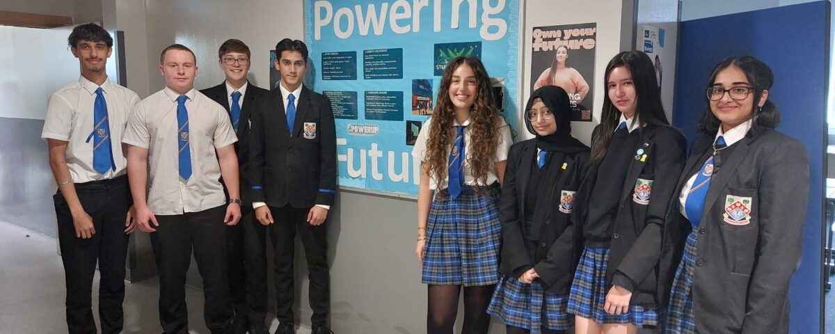 Eight high school students in uniform stand smiling in a hallway next to a blue bulletin board that reads “Powering our future.” Some wear blazers while others wear shirts and ties.