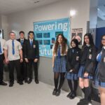 Eight high school students in uniform stand smiling in a hallway next to a blue bulletin board that reads “Powering our future.” Some wear blazers while others wear shirts and ties.